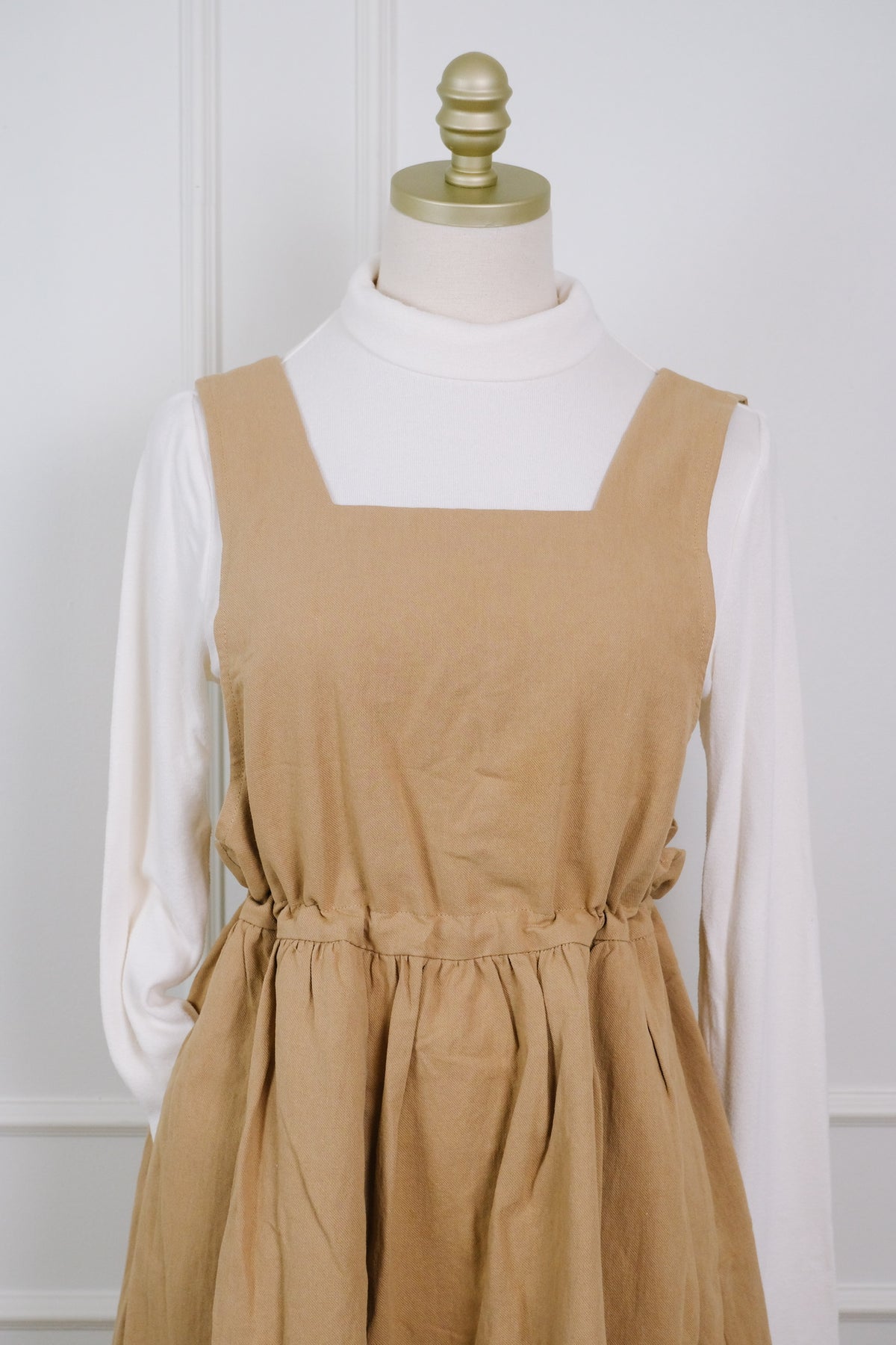 A tan linen pinafore dress with a square neckline and gathered waist, styled over a white mock neck long-sleeve shirt on a cream dress form with a gold finial.