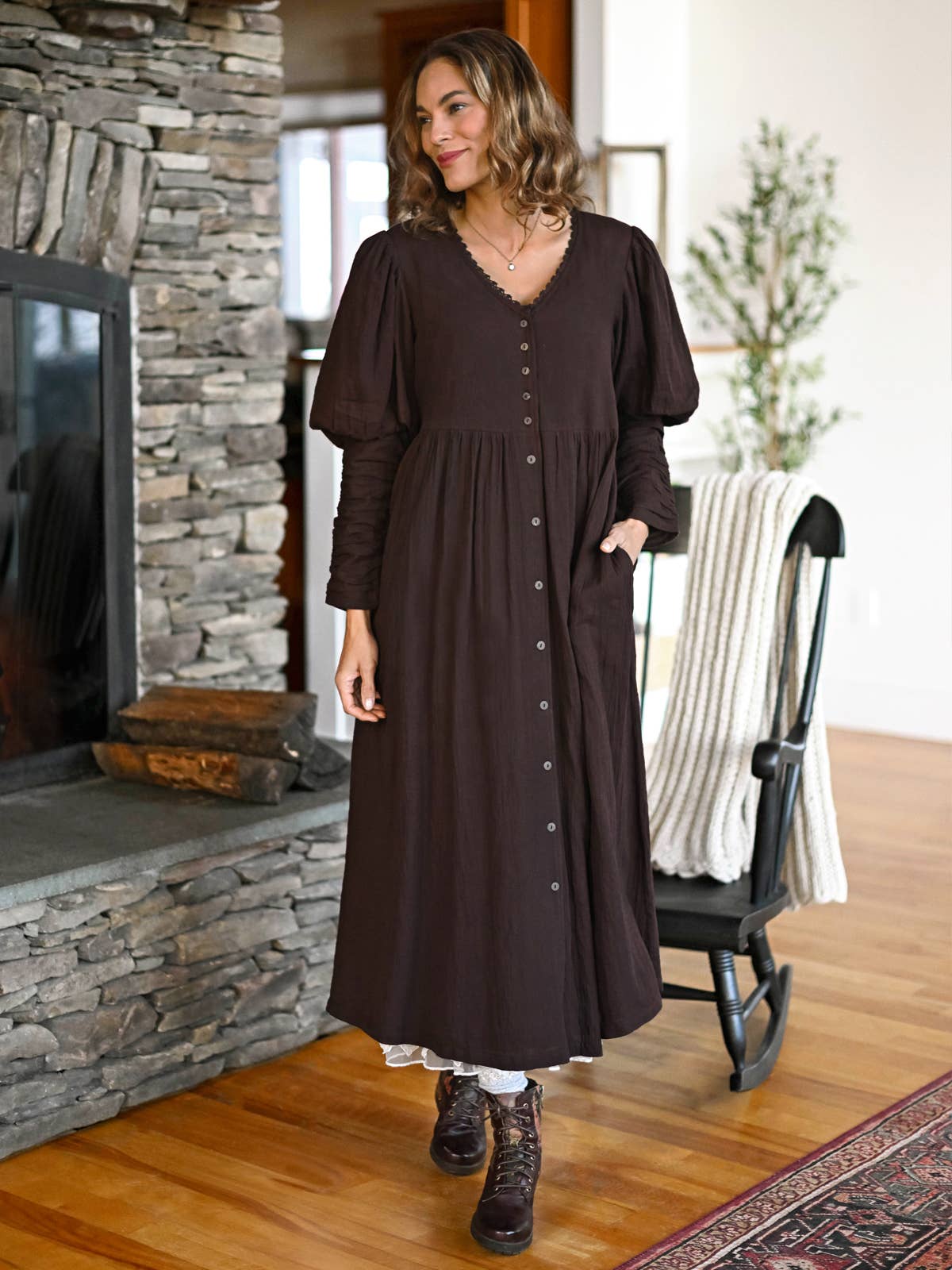 A woman wearing a dark brown long-sleeve midi dress with a button-down front and pockets, standing in a cozy room with a stone fireplace and wooden floor.