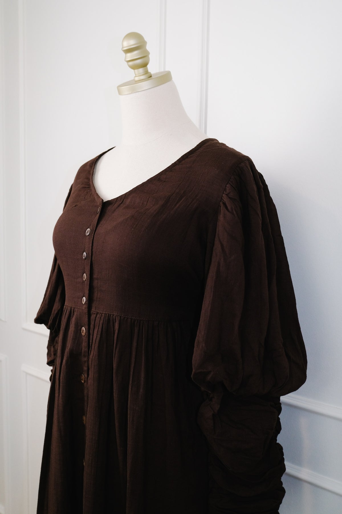 A dark brown long-sleeve button-front dress displayed on a cream mannequin torso against a white paneled wall.
