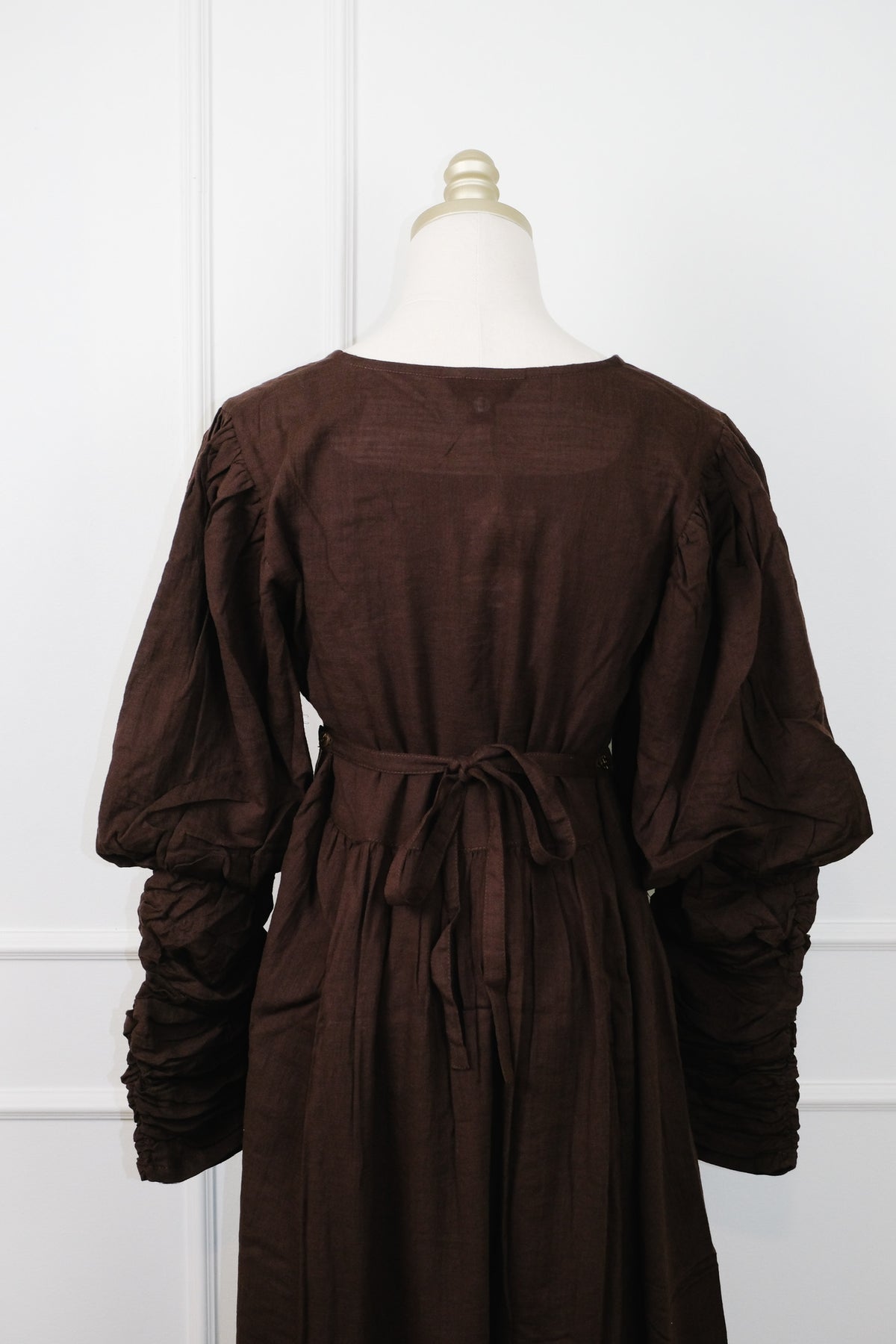 A dark brown long-sleeve dress displayed on a cream dress form mannequin against a white paneled wall. The dress features a tie waist, gathered sleeves, and a relaxed silhouette.
