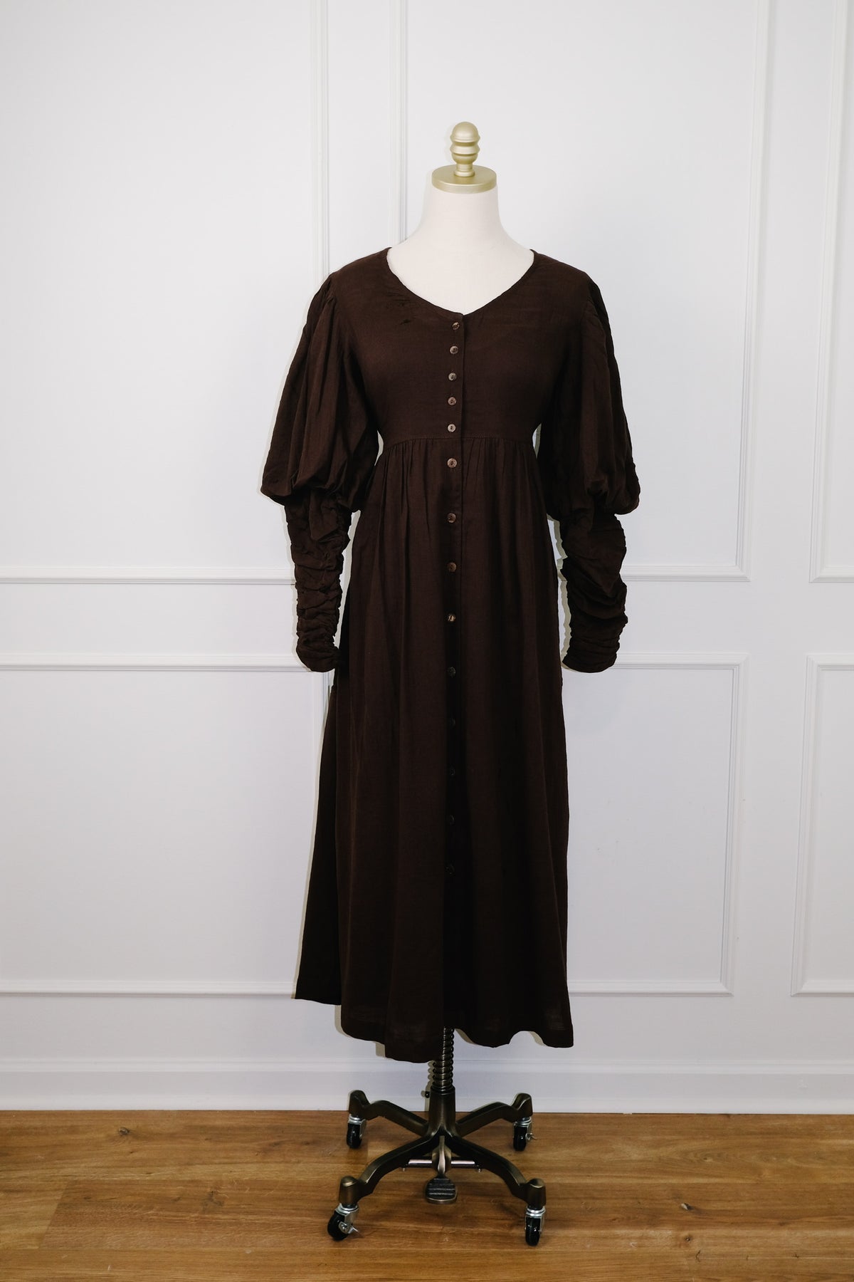 A dark brown midi dress with long puff sleeves and a button-down front, displayed on a mannequin stand against a white paneled wall and wooden floor.