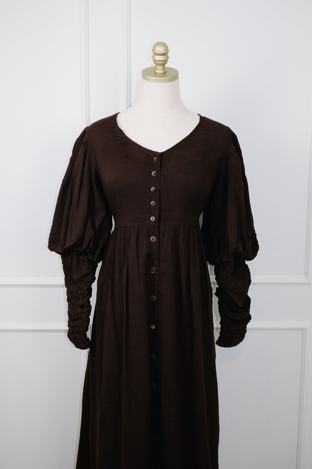 A dark brown long-sleeve button-front dress displayed on a cream mannequin torso against a white paneled wall.