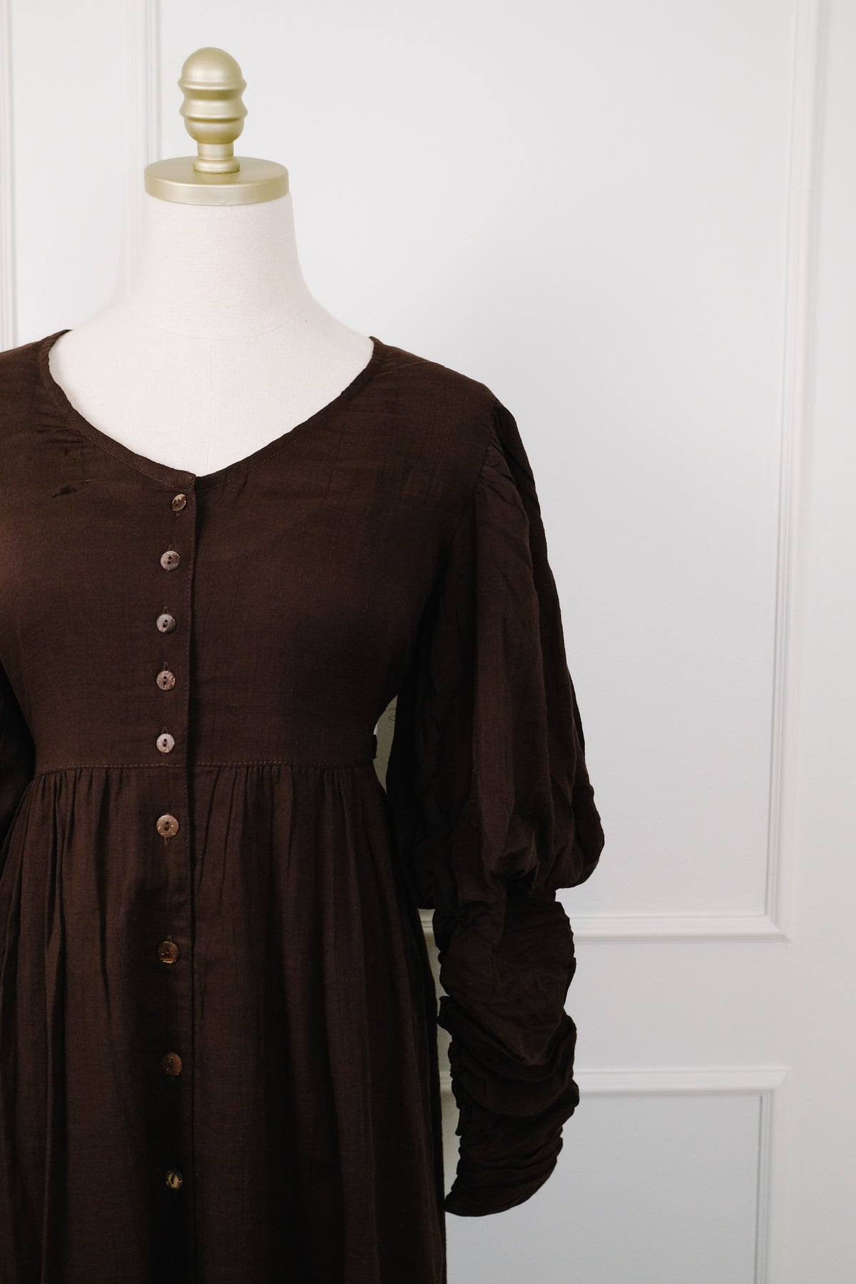 A dark brown long-sleeve dress displayed on a cream dress form with a gold finial, set against a white paneled wall.