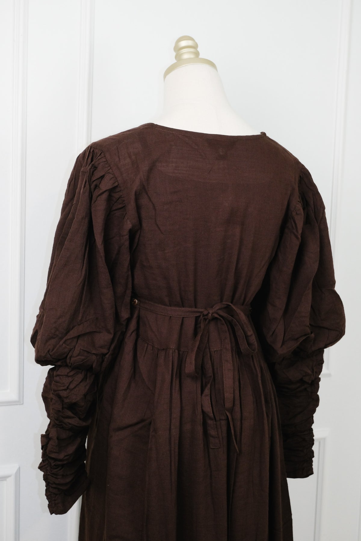 A dark brown linen dress displayed on a cream mannequin torso, showing the back view with gathered long sleeves and a tie waist belt.