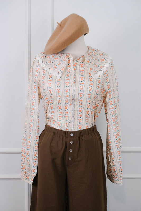A vintage-style long-sleeve blouse with a cream base and orange floral pattern, styled on a mannequin with a tan beret and brown pants against a white paneled wall.