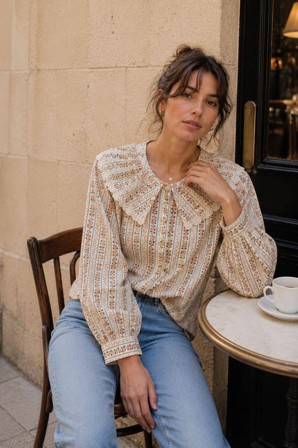 A woman sits on a wooden chair at a cafe table, wearing a cream-colored long-sleeve blouse with a dense multicolor geometric pattern and a large ruffled collar, paired with light blue jeans.