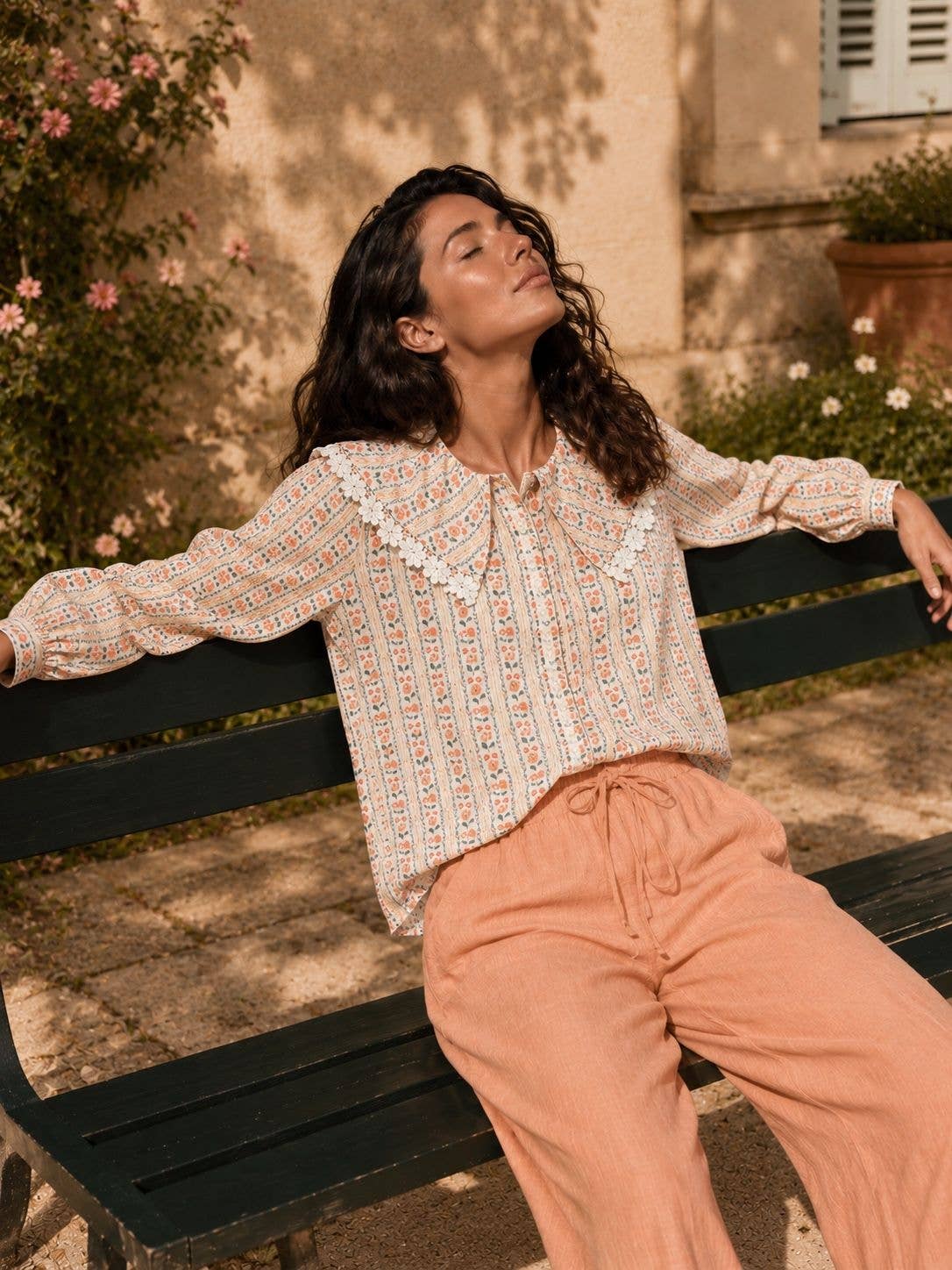 A woman lounges on a dark green park bench wearing a cream-colored long-sleeve blouse with a delicate multicolor floral pattern and a ruffled collar, paired with peach-colored linen pants.