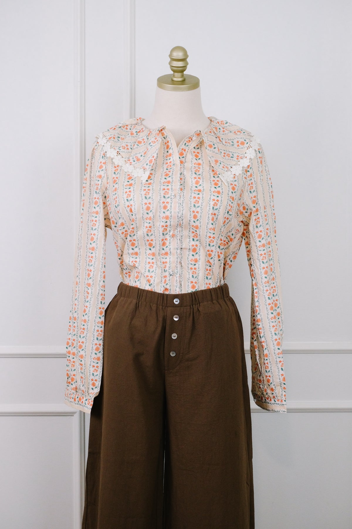 A long-sleeve blouse with a small orange and blue floral pattern on a cream background, styled on a mannequin with brown wide-leg trousers against a white paneled wall.