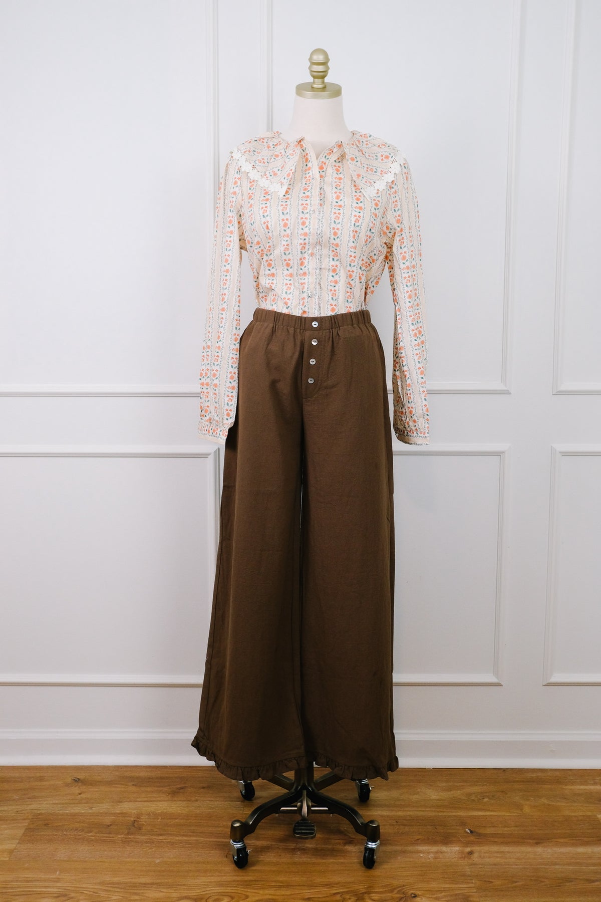 A vintage two-piece outfit displayed on a dress form, featuring a long-sleeve button-up blouse with a white base and orange geometric pattern, paired with high-waisted brown wide-leg trousers with a button fly.