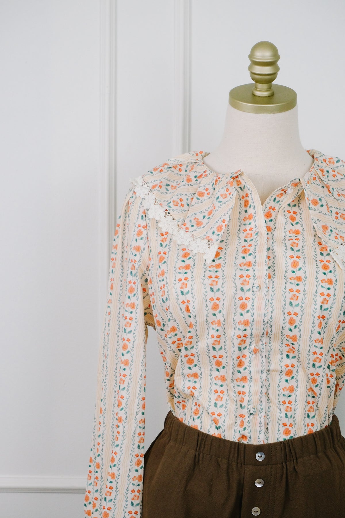 A long-sleeve blouse with a cream base and orange, green, and blue vertical floral stripes, styled on a dress form with dark brown pants against a white paneled wall.