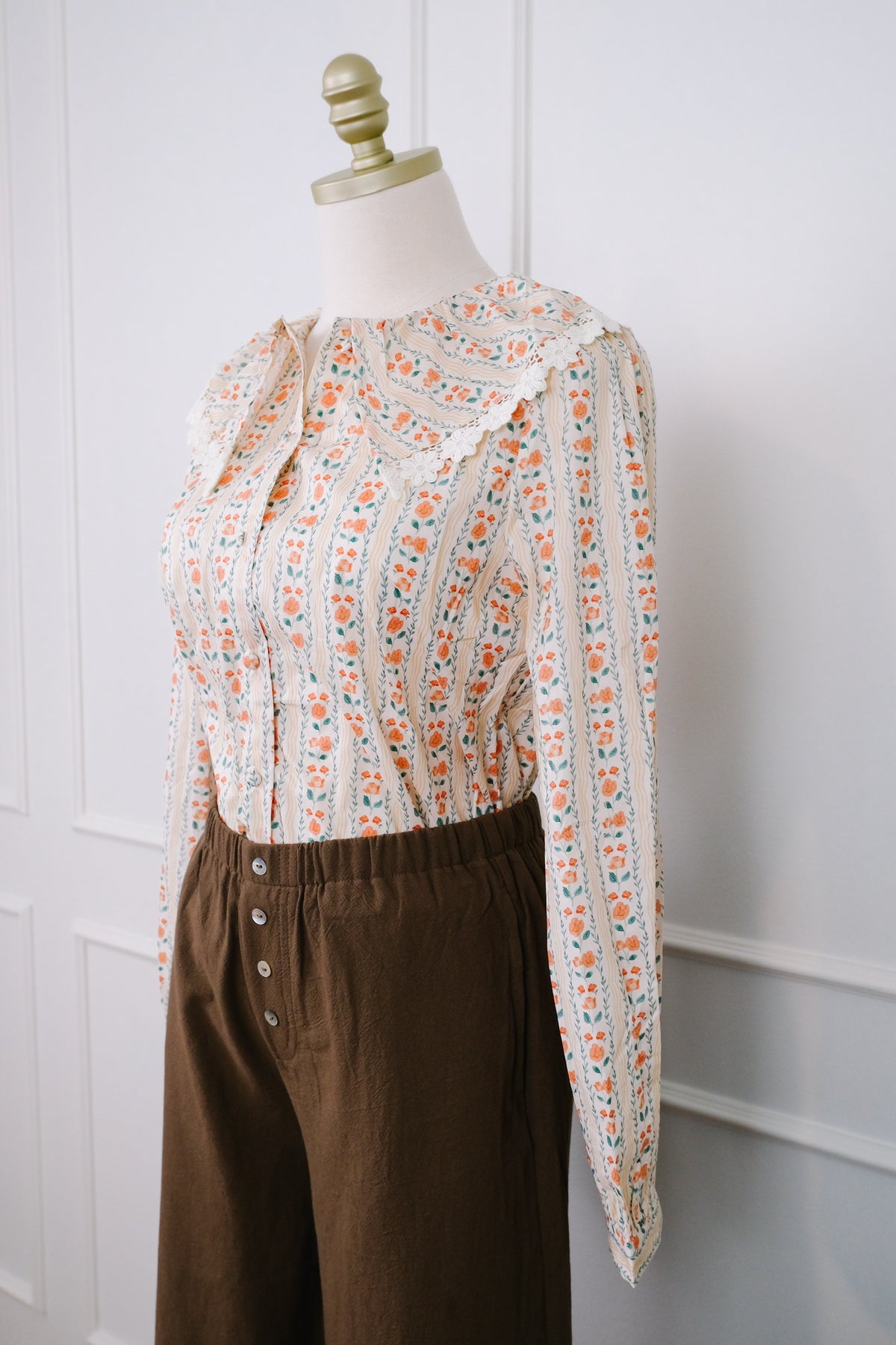 A long-sleeve blouse with a cream base and orange, green, and blue floral vertical stripes, styled on a mannequin with brown button-front trousers against a white paneled wall.