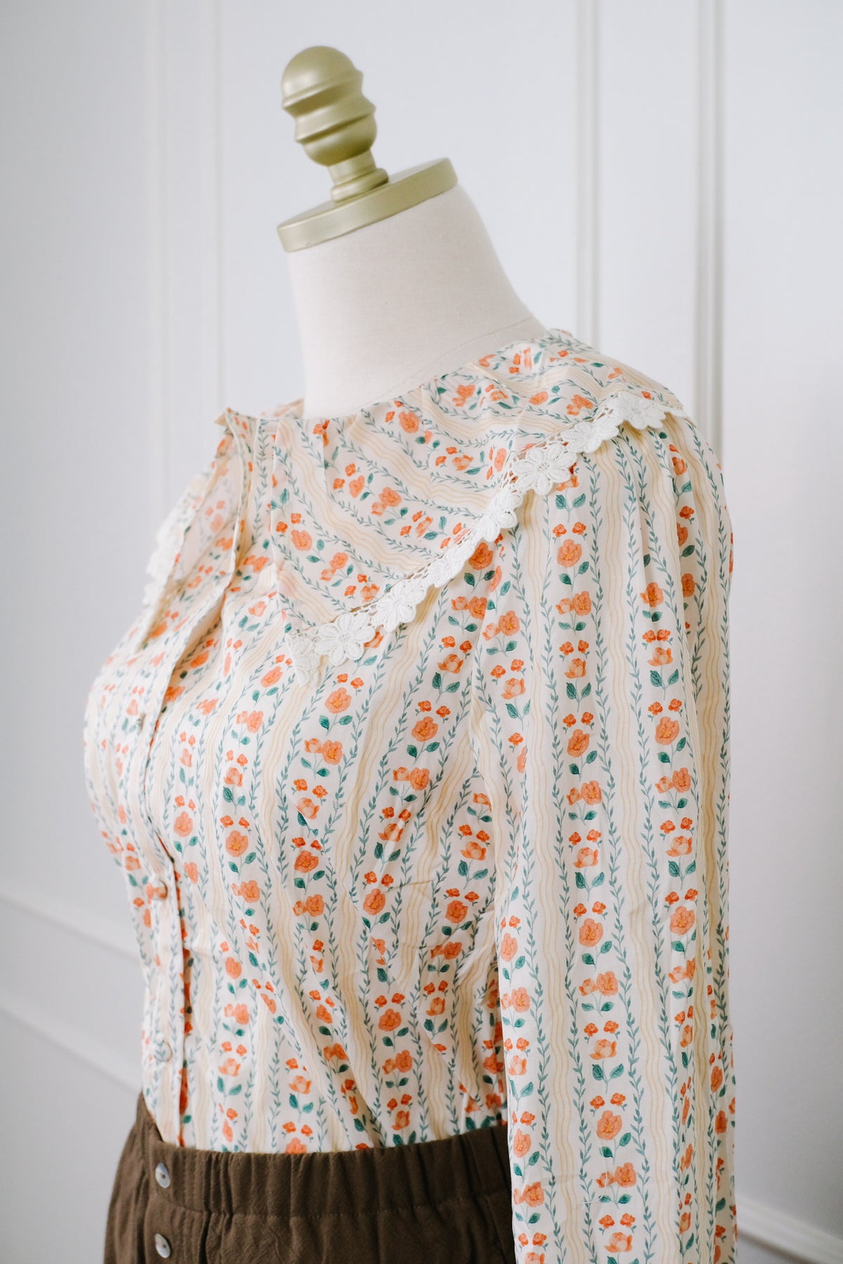 A long-sleeve blouse with a cream base and orange floral print, styled on a mannequin torso with dark brown pants against a white paneled wall.