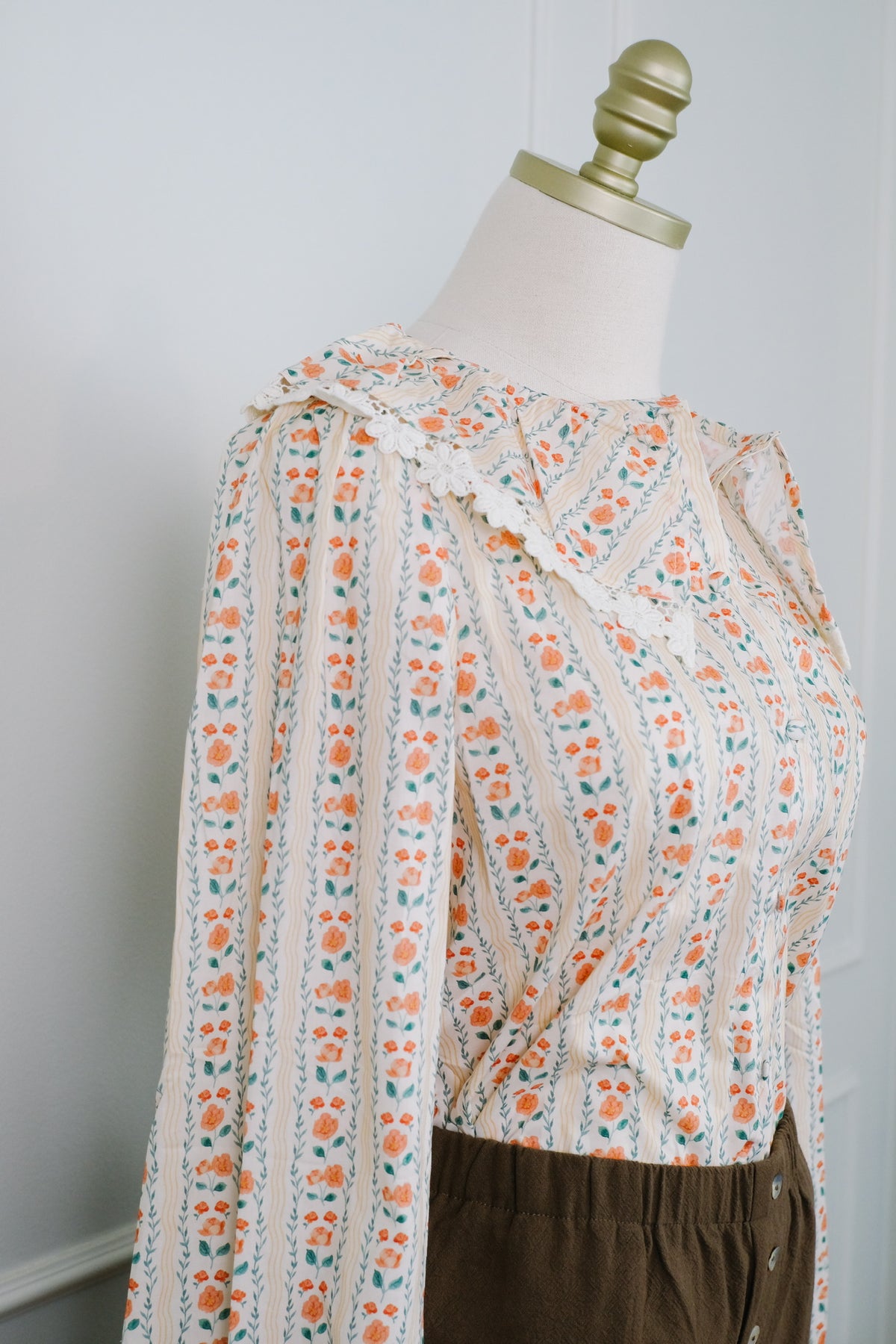 A cream-colored long-sleeve blouse with a vibrant orange and teal floral pattern, styled on a mannequin torso with dark brown pants against a light gray wall.