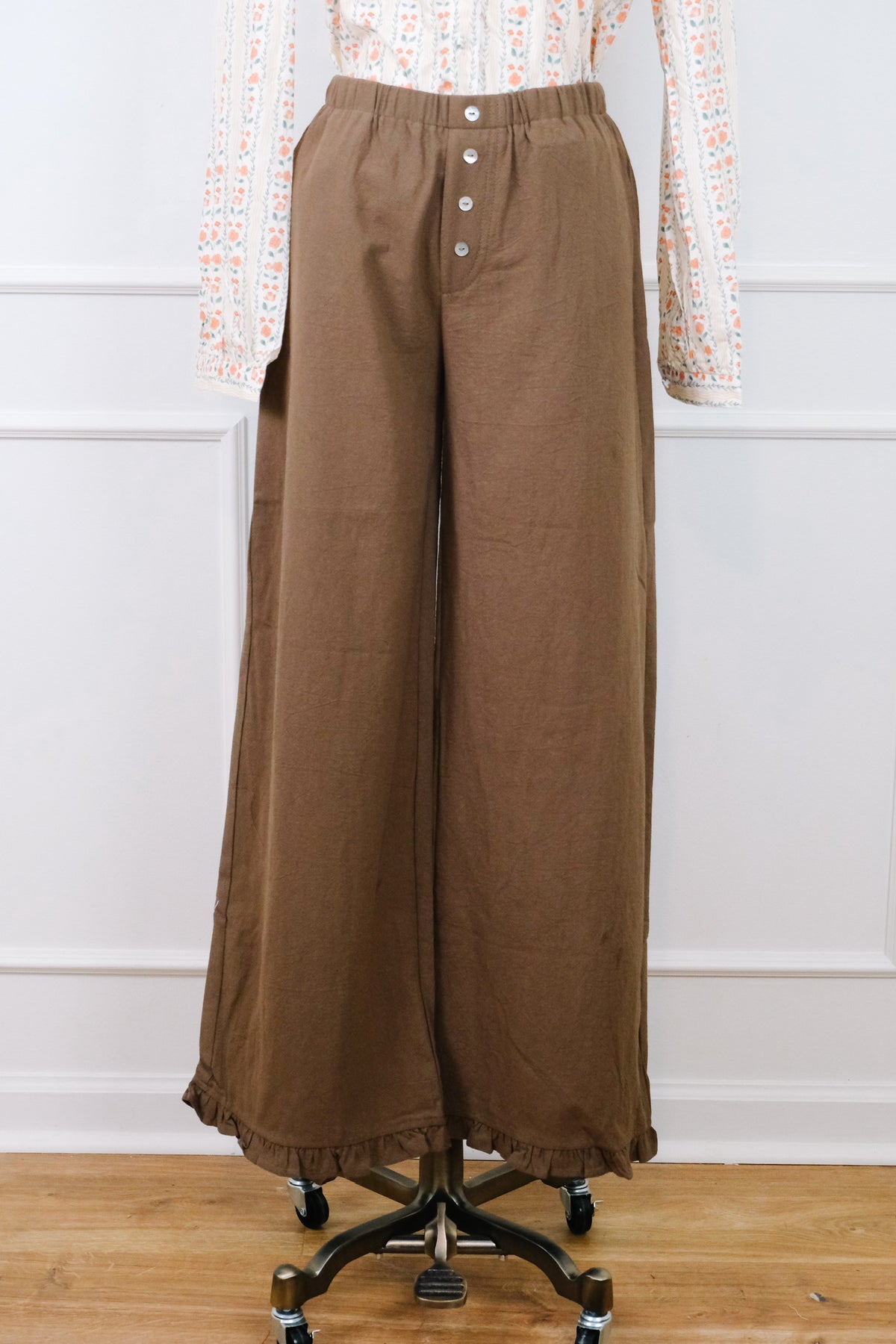 A pair of wide-leg brown pants displayed on a dress form mannequin, styled with a patterned long-sleeve top. The pants feature an elastic waistband with a button fly and a ruffled hem, set against a white paneled wall and light wood floor.