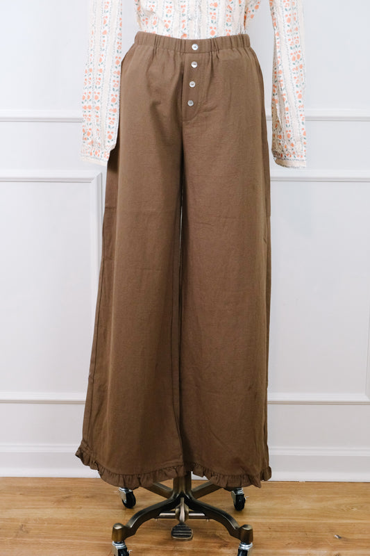 A pair of wide-leg brown pants displayed on a dress form mannequin, styled with a patterned long-sleeve top. The pants feature an elastic waistband with a button fly and a ruffled hem, set against a white paneled wall and light wood floor.