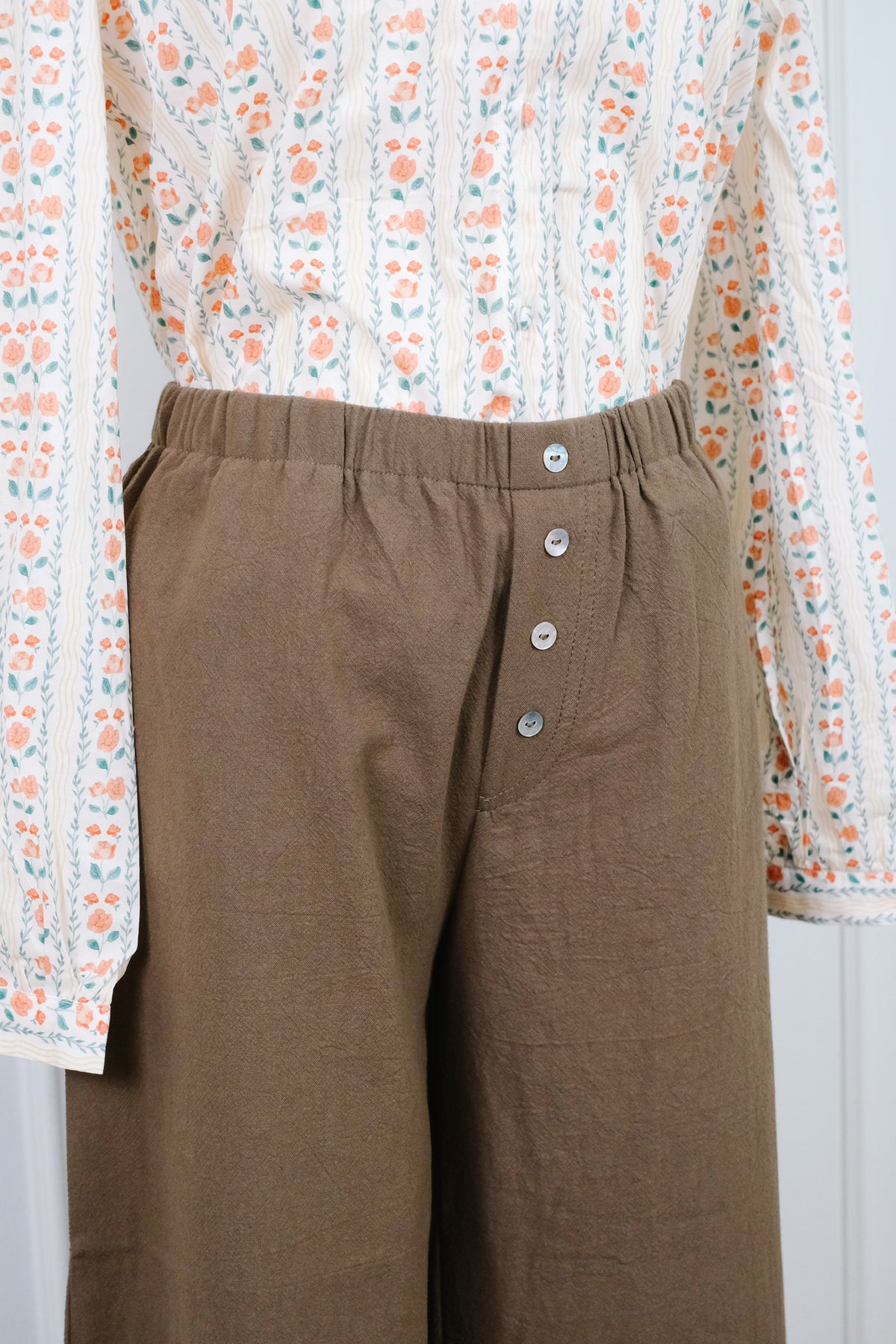 A close-up of a person wearing brown wide-leg trousers with a button fly and an off-white floral long-sleeve shirt.
