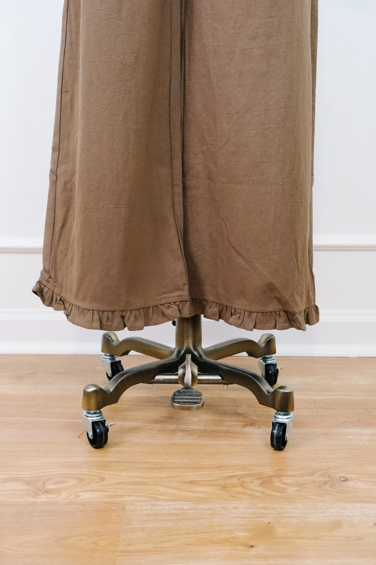 A brown ruffled fabric garment or drape hanging from a dark metal rolling stand with casters, shown against a white wall and light wood floor.