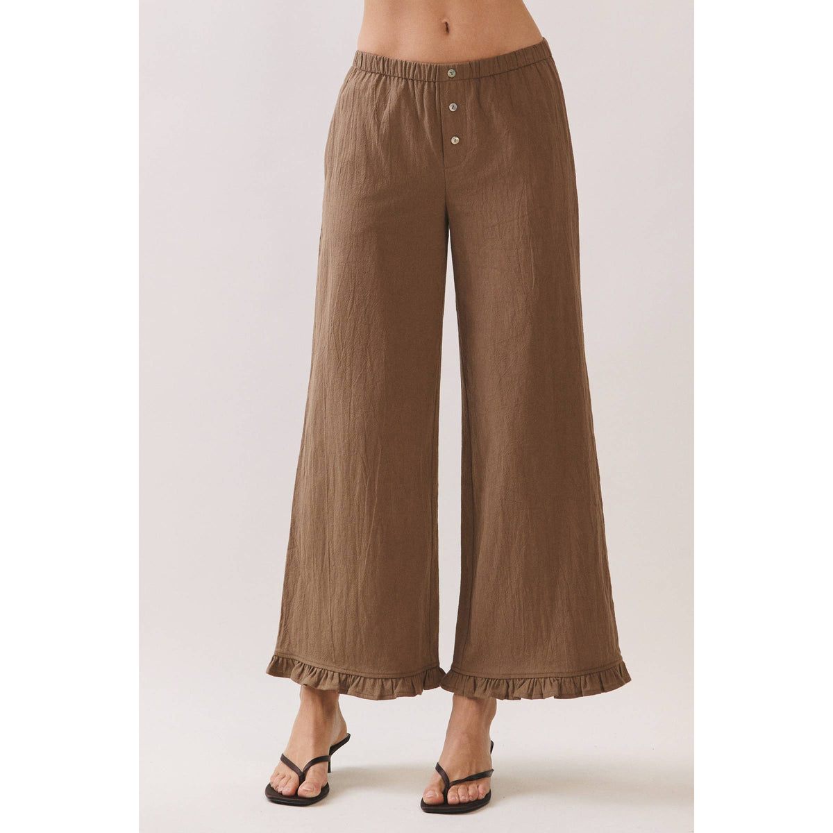 A model wearing brown wide-leg cropped pants with a buttoned waistband and ruffled hems, paired with black flat sandals, shown against a light beige studio background.