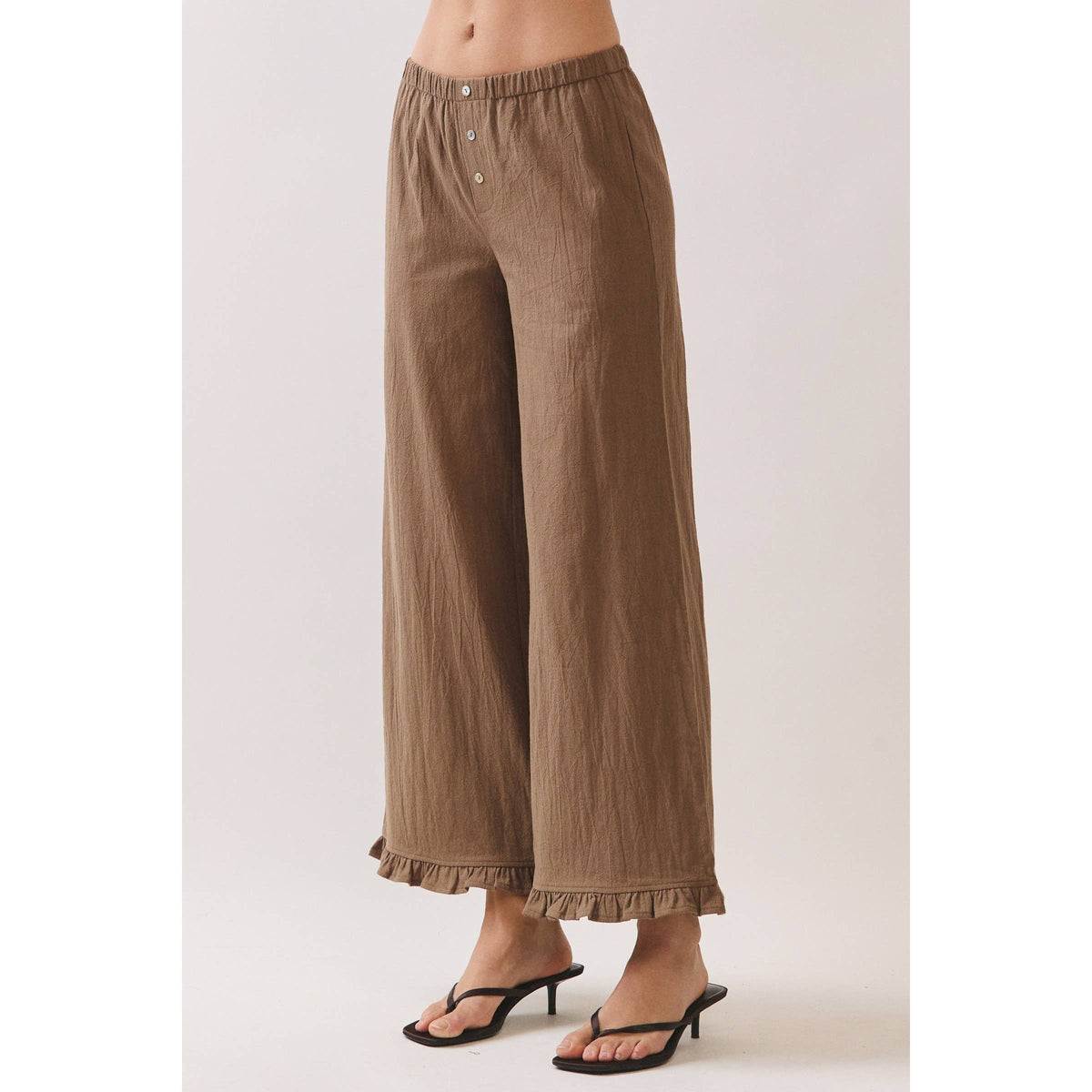 A model from the waist down wearing brown wide-leg cropped pants with a ruffled hem and button waistband, paired with black thong sandals against a light beige studio background.