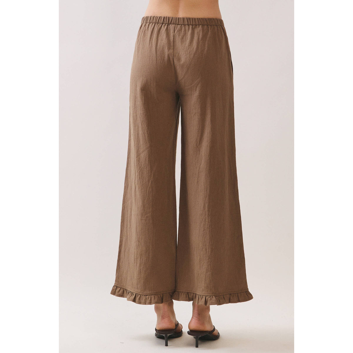 A person shown from the back wearing loose, wide-leg cropped pants in a warm taupe-brown color, paired with black heeled sandals against a plain light studio background.